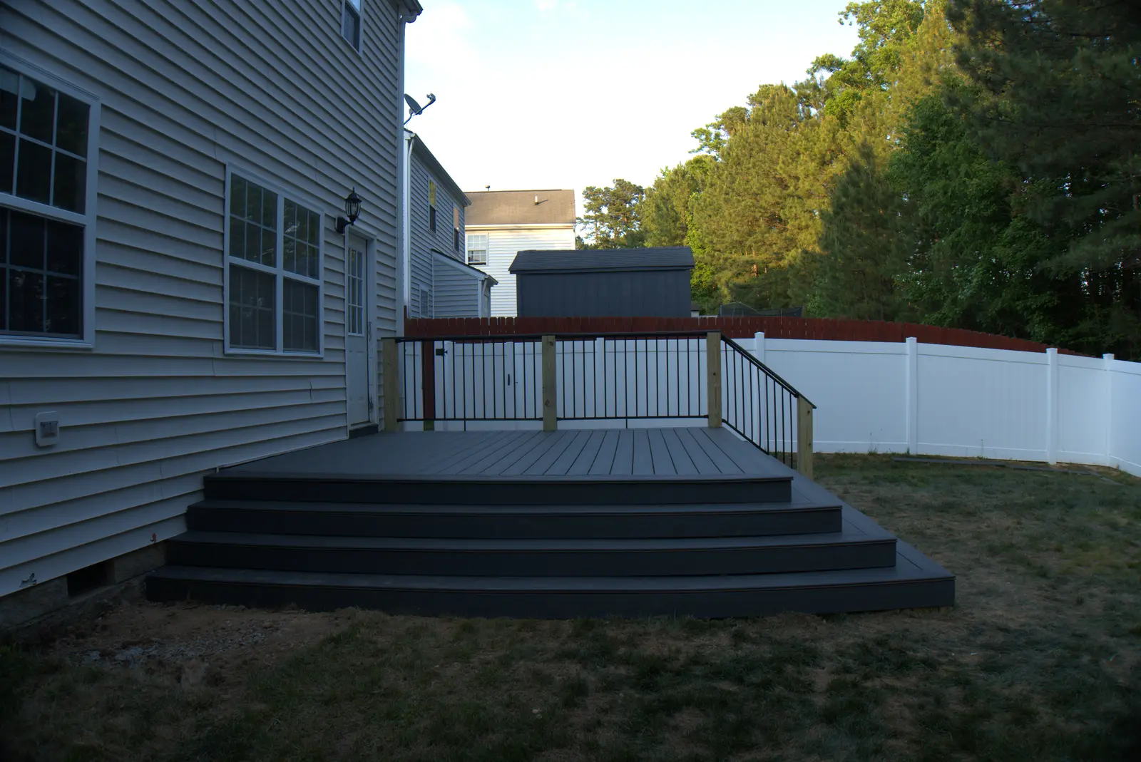 Grey composite deck with wide wraparound stairs and black railing behind a two story house