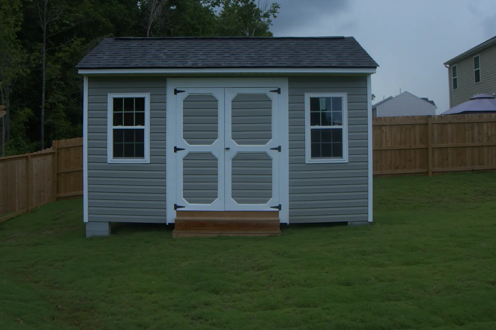 Grey vinyl storage building with white double doors and two windows in a fenced back yard