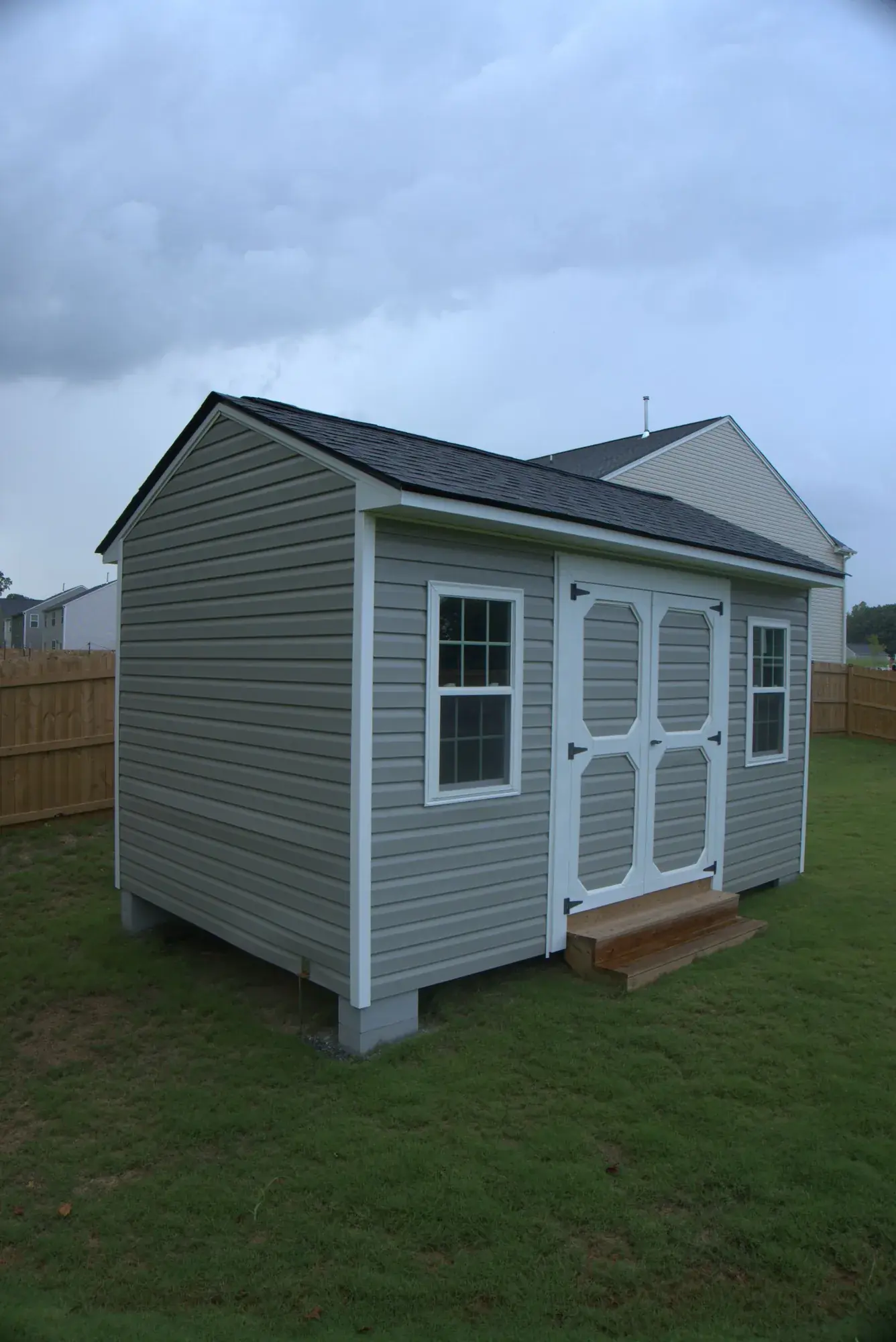 Finished grey vinyl storage building with white trim, double doors, two windows and a shingled roof, set on blocks in a fenced back yard