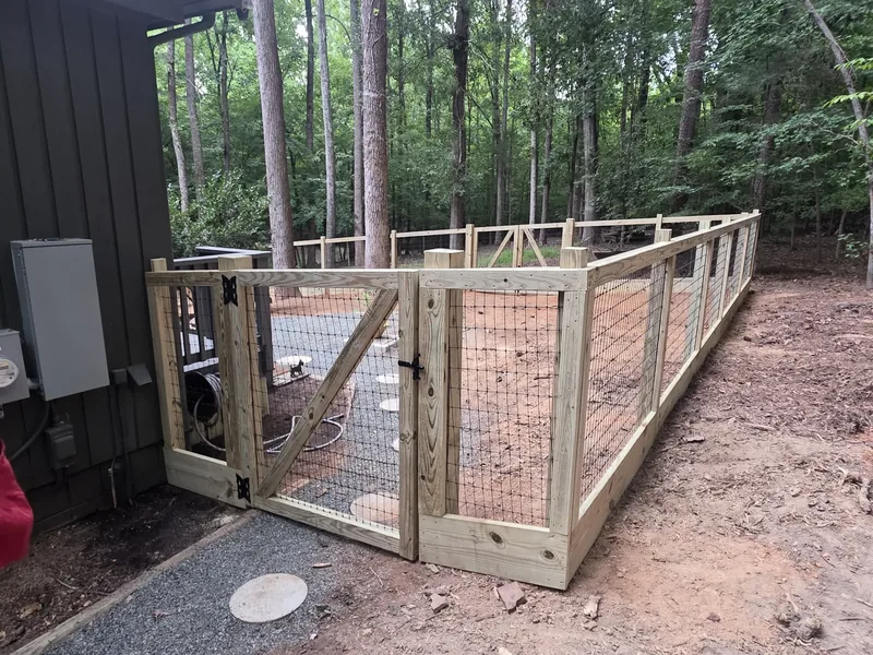 Finished wood-framed wire mesh fence with a braced gate beside a dark board-and-batten house